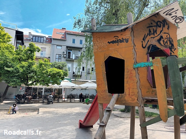 Une aire de jeux pour enfants dans le centre de Zagreb, avec une cabane au premier plan et une terrasse de café pour les parents en arrière-plan.