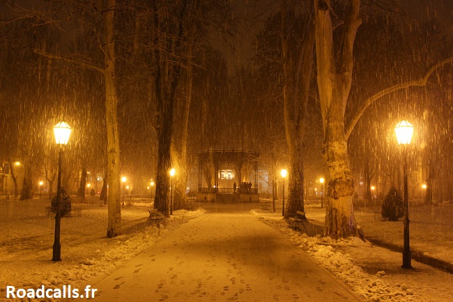 Le kiosque du parc de Zrinjevac dans le centre de Zagreb, en hiver, de nuit, sous une averse de neige, avec les réverbères diffusant une lumière jaune triste, et le sol tapissé de neige avec des traces de pas.