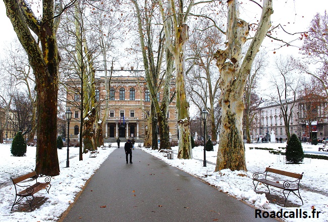 Une des allées du parc de Zrinjevac sous la neige, avec les bancs, les immenses platanes, et l'atmosphère hivernale de Zagreb.
