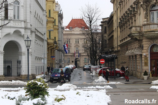 Une rue de Zagreb en hiver, partiellement déneigée, avec des voitures garées et des piétons qui traversent.