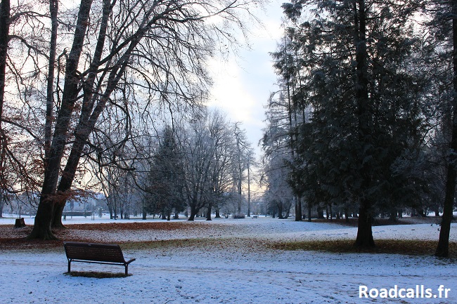 Un banc vide dans un parc arboré, le sol tapissé de feuilles mortes et de neige, sous un ciel gris où perce une pâle lumière jaune de fin de journée, à proximité de Zagreb, au coeur de l'hiver croate.