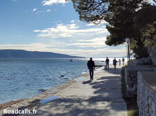 Promenade de bord de mer à Cres, en fin de journée, sous le ciel bleu légèrement voilé, avec des silhouettes de promeneurs en contre-jour.