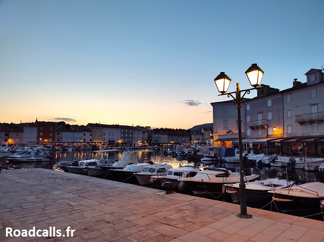 Le port de Cres sur l'île de Cres, en Croatie, au moment du coucher de soleil. Un lampadaire est allumé, les bateaux dorment sur le port, et les enseignes des restos et des cafés sont allumées.