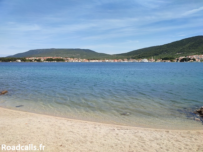 Petite plage de cailloux au premier plan, puis la mer avec au loin la ville de Cres et ses toits orange, puis ses collines.