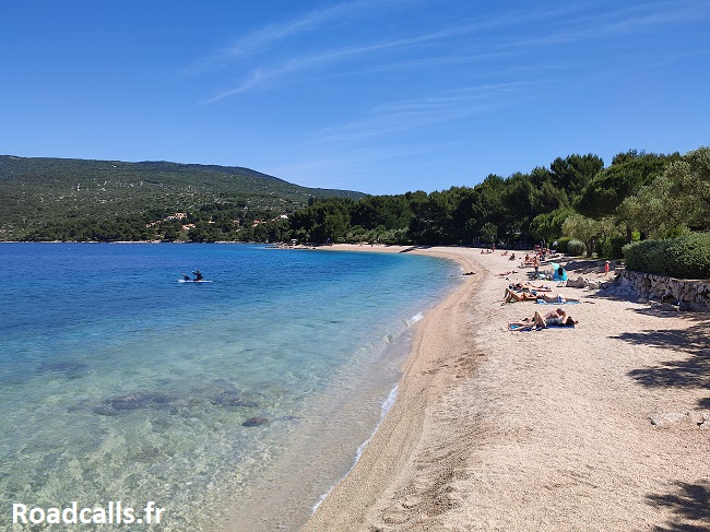 La plage de Kovacine et ses eaux limpides, sous un ciel bleu : les vacanciers lézardent au soleil.